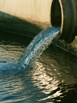 Industrial pipe discharge pouring clear water into retention basin under warm sunlight, creating rippling reflections beside concrete wall, representing civil engineering infrastructure