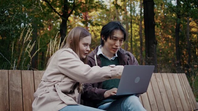 Couple reviews laptop together on wooden bench in autumn park