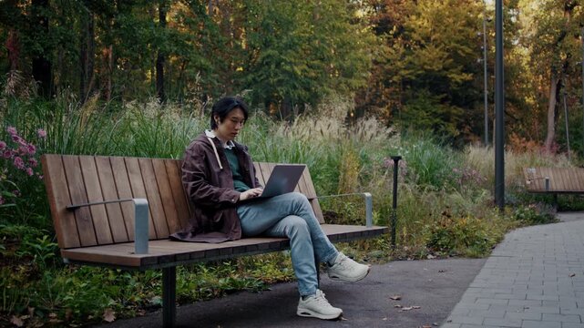 Asian man works on laptop while sitting on autumn park bench