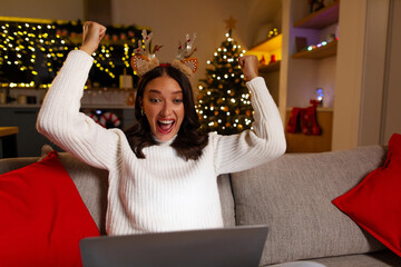 Overjoyed woman in reindeer headband reacting happily to news on laptop during Christmas at home, copy space © Home-stock
