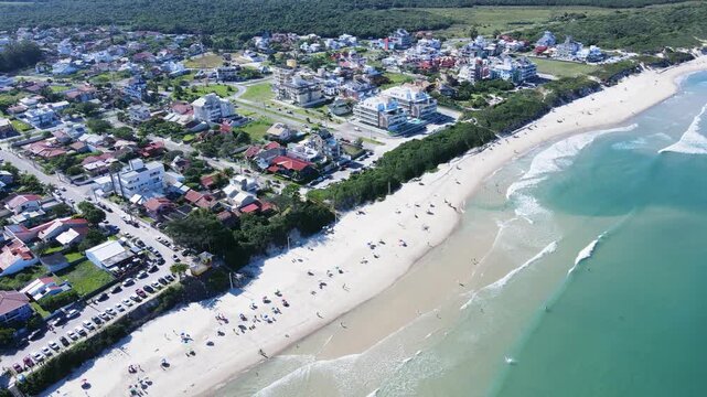 Drone aerial panoramic view of Praia dos Acores residential coastal neighborhood in Florianopolis, Brazil, with tourists on white sand beach, modern properties, and turquoise ocean waves
