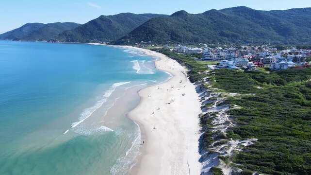 Drone aerial panoramic view of Praia dos Acores residential coastal town looking towards Solidao beach and Saquinho cape in Florianopolis, Brazil, with turquoise ocean and green mountains
