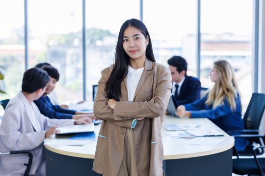 Portrait of smiling young Asian businesswoman standing with arms crossed a diverse group of business colleagues is working and collaborating around a meeting in modern office meeting room.