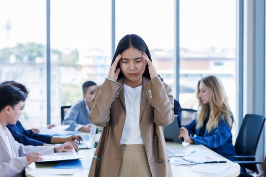 Stressed young Asian businesswoman holding her head with hands a diverse group of business colleagues is working and collaborating around a meeting in modern office meeting room.