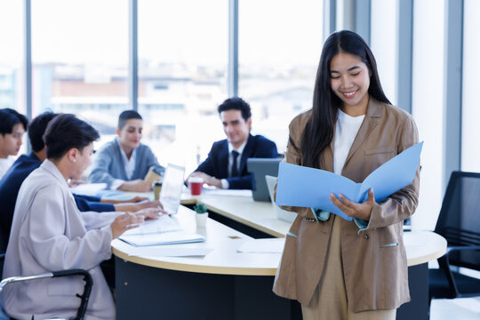 Portrait of smiling young Asian businesswoman standing with arms crossed a diverse group of business colleagues is working and collaborating around a meeting in modern office meeting room.