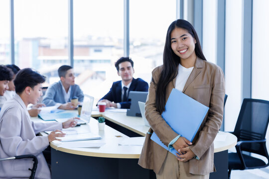 Portrait of smiling young Asian businesswoman standing with arms crossed a diverse group of business colleagues is working and collaborating around a meeting in modern office meeting room.