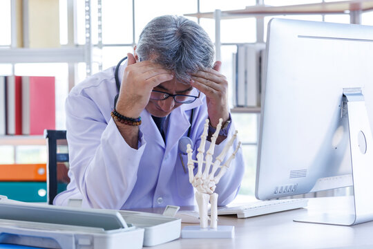 Stressed and exhausted senior male doctor wearing a lab coat and stethoscope, leaning on his desk with hand on forehead.fatigue, headache, or medical crisis while working computer monitor at a clinic.