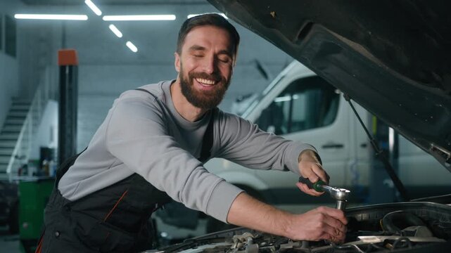 Smiling Caucasian man mechanic checking engine fault under open hood garage male technician using spanner inspecting car problem happy repair worker service station doing mechanical diagnosis indoors