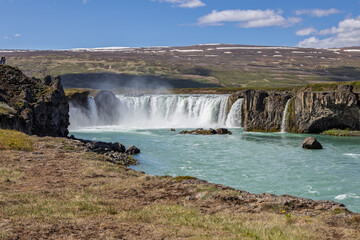 White cascades stretch between dark rocky banks at Godafoss in North Iceland, sending mist over the turquoise Skjalfandafljot River beneath hills with lingering snow. © Artem