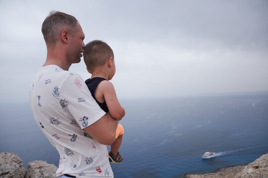 A man carries a young boy at a coastal viewpoint as both look toward a boat below, connecting family attention with a wide sea view