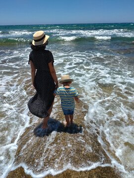 Viewed from behind, an adult guides a child through foamy water at the shoreline, highlighting reassurance during a shared seaside experience