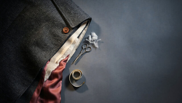 Gray herringbone wool tweed coat with brown button and burgundy satin lining beside vintage scissors and measuring tape on a dark background for tailoring and bespoke fashion