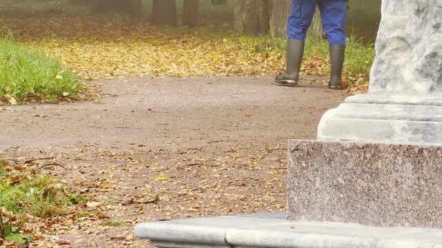 A park worker cleans a leaf-strewn path near a stone pedestal in Catherine Park, Pushkin, Saint Petersburg. The misty autumn morning evokes a calm and peaceful feeling.