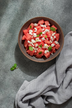 Salad of fresh watermelon, feta cheese and mint leaves in a bowl on a grey rustic background with sunlight, top view, vertical