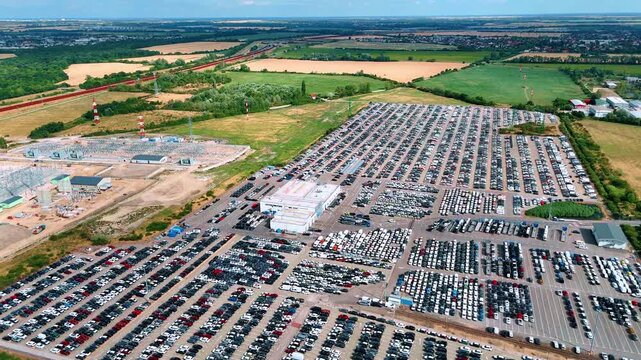 A vehicle storage complex stretches toward surrounding fields. Dense rows of cars flank paved aisles beside an industrial construction area.