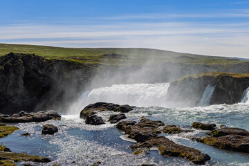 White water surges over rocky ledges on the Skjalfandafljot River near Godafoss, North Iceland, as sunlit spray rises above the cascades and moss covers foreground boulders. © Artem