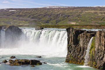 White cascades plunge over the dark cliffs of Godafoss in North Iceland, filling the turquoise river with foam and spray beneath sunlit green hills streaked with snow. © Artem