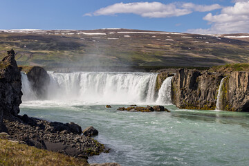 White cascades at Godafoss send spray over the Skjalfandafljot River, framed by dark rock, moss and a snow-streaked hillside under a blue summer sky in North Iceland. © Artem