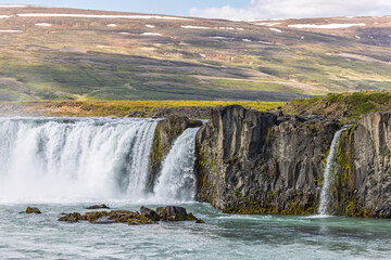 Powerful white cascades spill over dark basalt cliffs at Godafoss in North Iceland. Turquoise river currents swirl around rocks below hills streaked with lingering snow. © Artem