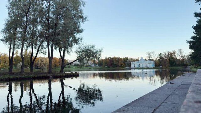 The light blue Grotto pavilion stands gracefully across the calm pond. Tall trees cast tranquil reflections on serene waters. This peaceful landscape shows Catherine Park, Pushkin, Saint Petersburg.