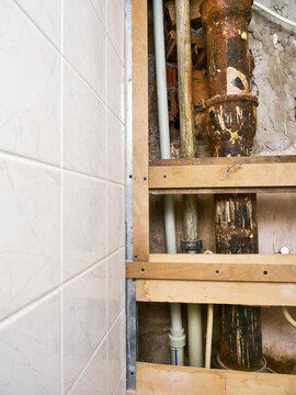 Exposed plumbing and wooden framing in tiled bathroom wall. Rusty pipes and exposed fixtures show renovation progress. Brown water filter attached to wall below frame