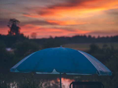 Blue beach umbrella at sunset near water. Sky glowing orange and red. Trees and grass in background. Calm atmosphere, peaceful evening scene. No animals or plants identified