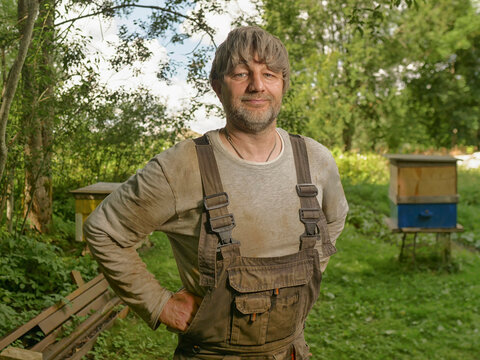 Man in his 40s or 50s in dirty over rolls standing close to bee hives. Green farmland with trees in the background. Farmer portrait in natural scenery. Working hard on a land theme. Calm and relaxed