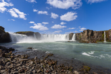 Godafoss stretches across a rocky ledge as white cascades churn the Skjalfandafljot River. A low viewpoint frames rough shoreline stones beneath a blue summer sky in North Iceland. © Artem