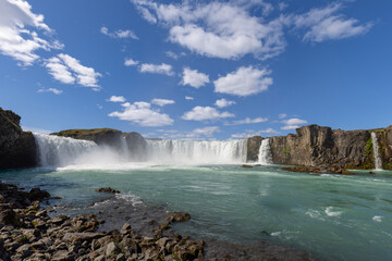 A low view across turquoise river water reveals the broad horseshoe of Godafoss in North Iceland, with foaming cascades, drifting spray and jagged rocks along the foreground shore. © Artem