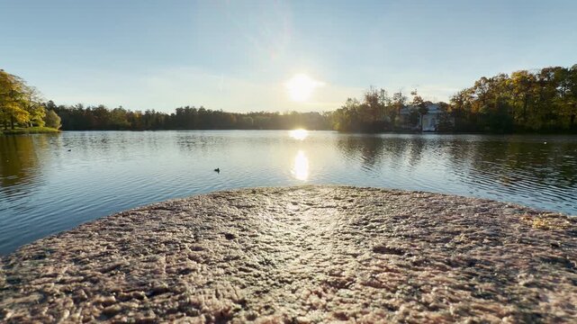 Bright sunlight glimmers over calm lake waters in Catherine Park, Pushkin, Saint Petersburg. A textured stone ledge leads into a serene, tranquil autumn horizon.
