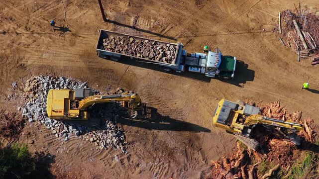 Two yellow crawler excavators work a land clearing site, one swinging its bucket of rocks into a dump trailer, the other in tree stumps. Workers in hi-vis vests, long shadows, sunny day.