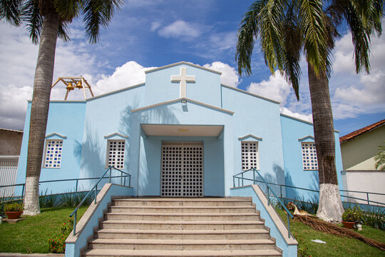 Vista frontal da Par&oacute;quia Nossa Senhora da Guia com dois coqueiros na frente e c&eacute;u nublado ao fundo.