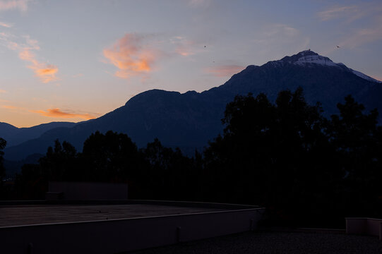 Soft orange clouds glow above a dark mountain silhouette and treeline at dusk, with a snowcapped peak and flat rooftop in the foreground creating a calm, moody evening landscape scene.
