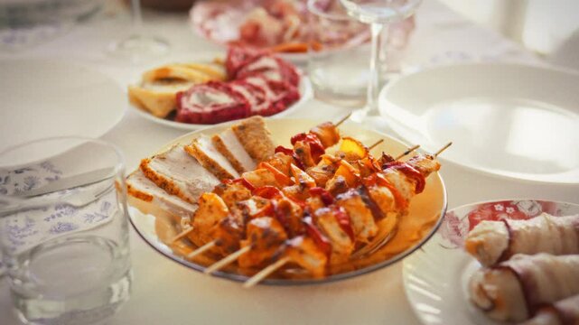 Anniversary lunch table in soft sunlight, close-up of sparkling empty glasses and white plates with sliced jamon and meat on skewers, a bright home celebration of a special family date