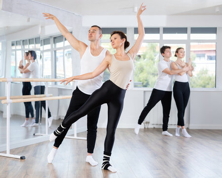 Pretty female ballerina and male partner practicing ballet arabesque pose during training with group active people in light dancing hall