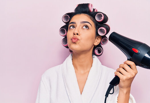 Young woman with hair rollers in white bathrobe holding black hair dryer whistling and looking up against pink wall