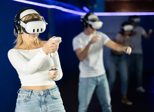 In darkened hall of gaming club, girl in VR helmet takes part in group game. Player overcomes obstacles and reaches goal in parallel reality. Imaginary fantastic world .