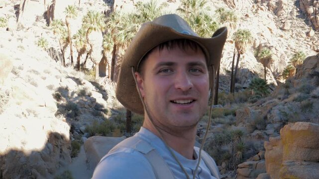 Cheerful male hiker in a cowboy hat filming a travel vlog while trekking through a rocky desert canyon, with wild palm trees, an oasis and sunny wilderness scenery