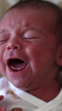 A three-week-old newborn baby cries while lying at home. The infant moves its arms and legs and fusses. Close-up shots of the baby's face and body during crying.