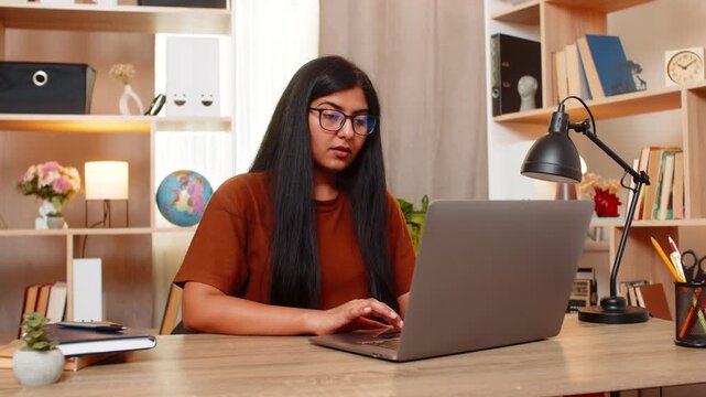 Indian woman on home table views offers on laptop and compares prices before choosing a product online. Arabian girl studies deals on screen while searching for a suitable purchase from home indoors.