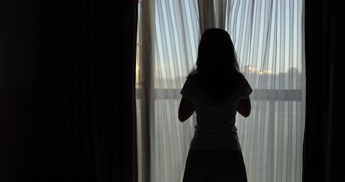 Woman looking through a balcony window, and closing curtain in apartment room	