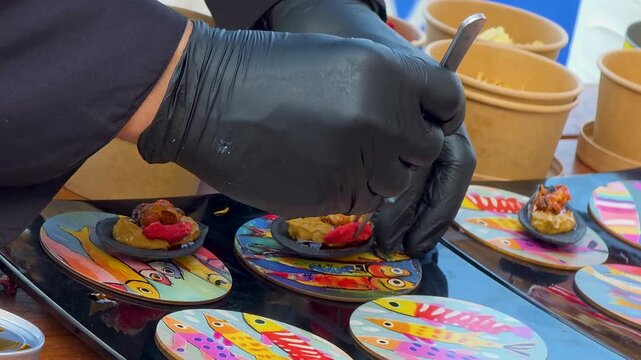 Chef in black gloves using tweezers for delicate plating of gourmet tapas on artistic coasters at a high cuisine gastronomy festival, close-up.