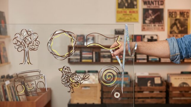 Hand pulling iridescent cassette tape ribbon on glass board in vintage record shop with tape art illustrations of tree, songbird, boombox, spiral, and banner for Lunar New Year