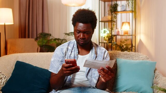 African American young man on home sofa holds bill checks amount and looks concerned with smartphone. Black guy on home sofa feels surprise and starts counting monthly expenses carefully in phone app.