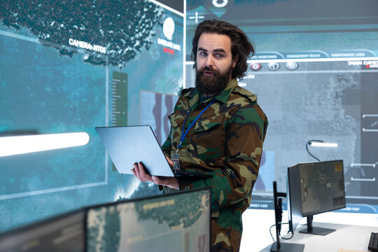 Military personnel, laptop and digital surveillance as uniformed man carries open computer near maritime map displays. Communication, planning and intelligence review support work in command center.
