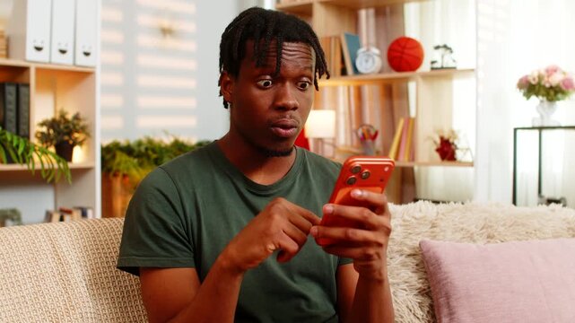 African American young man on home sofa reading shocking smartphone news with open mouth surprise in cozy living room. Black guy on home sofa reacting to unexpected phone message with amazed emotion.