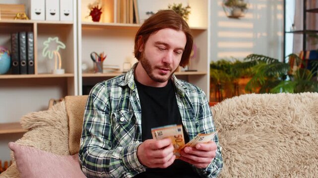 Young man on home sofa counts cash notes with proud smile and checks earnings with satisfaction. Adult guy on home sofa shows banknotes with pleased mood and enjoys success during calm indoor leisure.