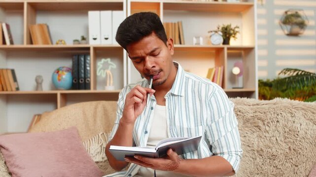 Indian man on home sofa writes notes in notebook and bites pen while thinking about ideas in living room. Arabian guy on home sofa plans work or tasks with focus during indoor evening break at home.