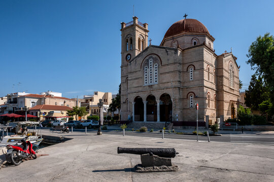 Church of Panagitsa (The Presentation of the Virgin) features local limestone architecture, a prominent dome, and a bell tower, with a historic cannon displayed in the foreground plaza. Aegina, Greece