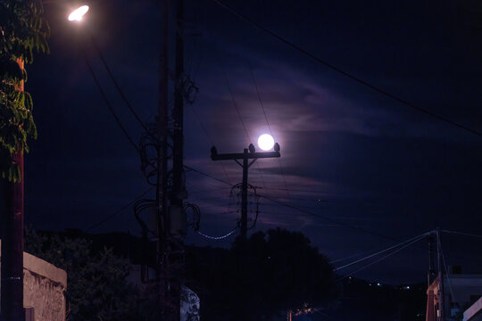 A bright full moon shines through wispy night clouds directly behind an electricity pole with power lines, illuminating the dark street and rooftops of a coastal village on Aegina Island, Greece.